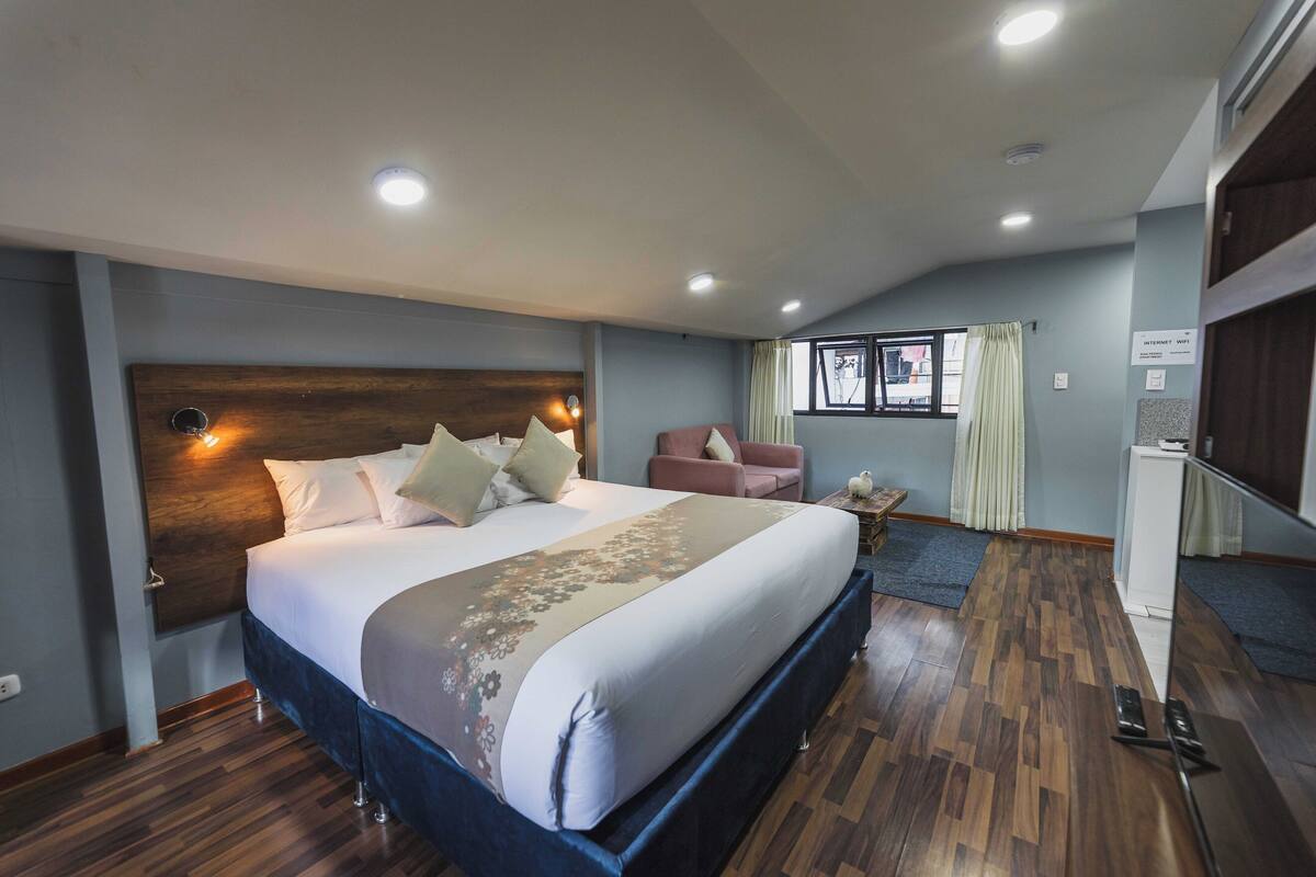 A hotel room with a large bed, a small seating area, a window with curtains, and a refrigerator.