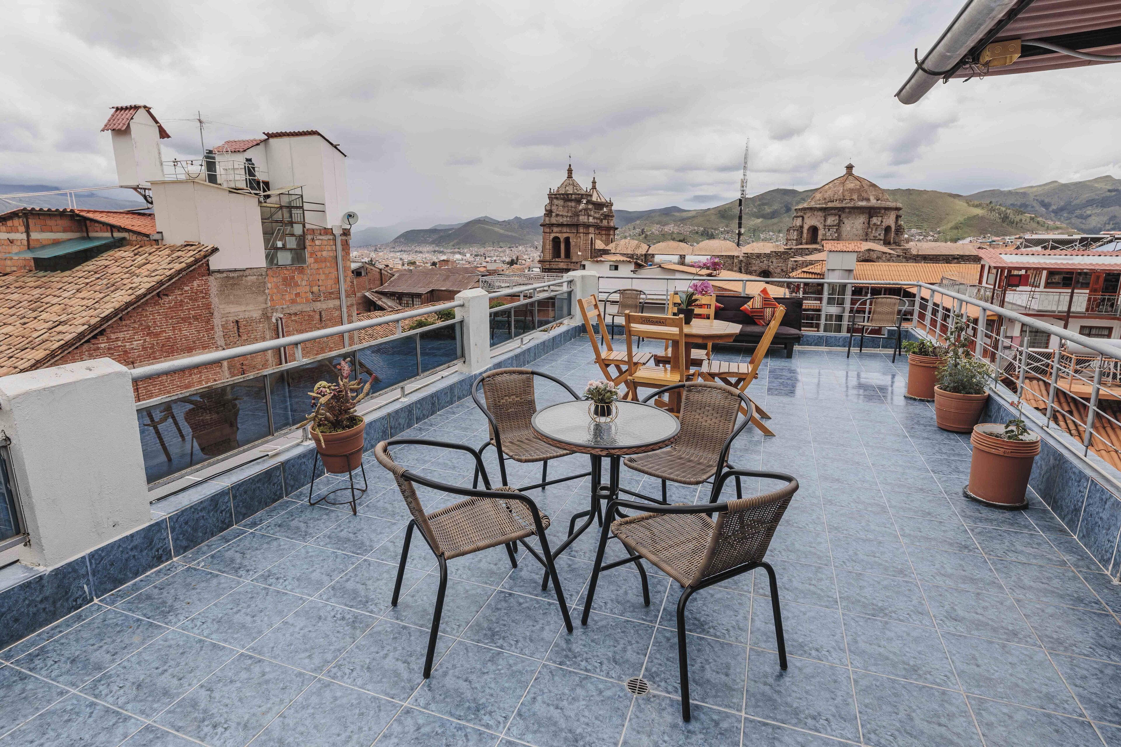A rooftop terrace with a round table and wicker chairs, overlooking a cityscape with historical buildings and mountains.