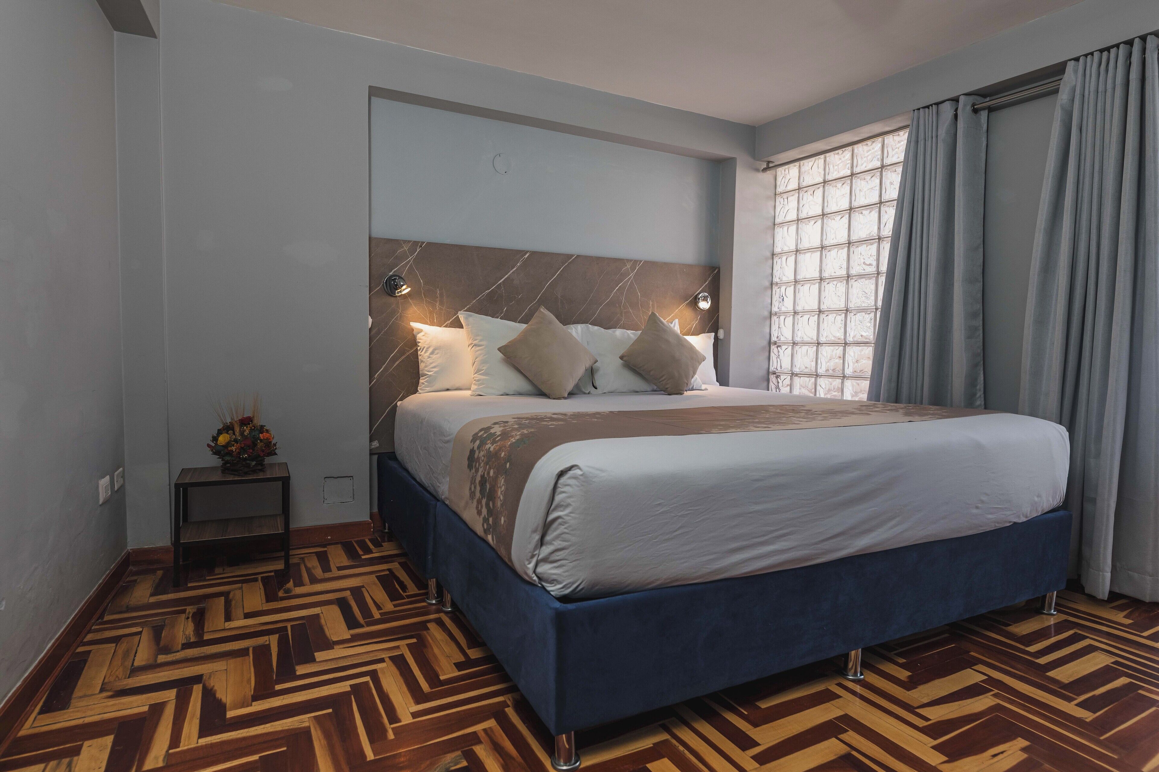 A modern hotel room with a large bed, a wooden parquet floor, a marble headboard, and a window with curtains.