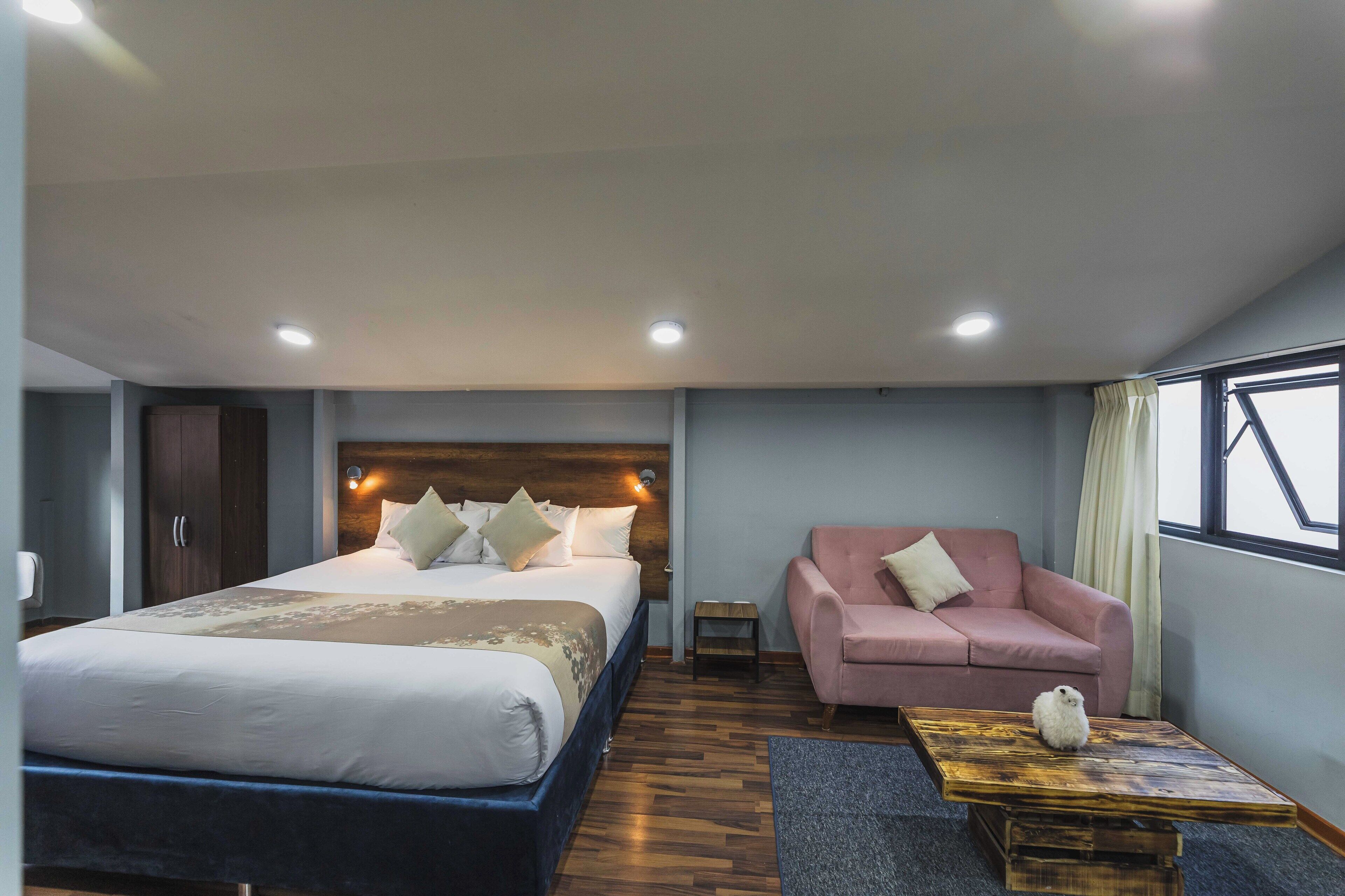 Una habitación de hotel con una cama grande, un sofá rosa, una mesita pequeña y una ventana con cortinas.