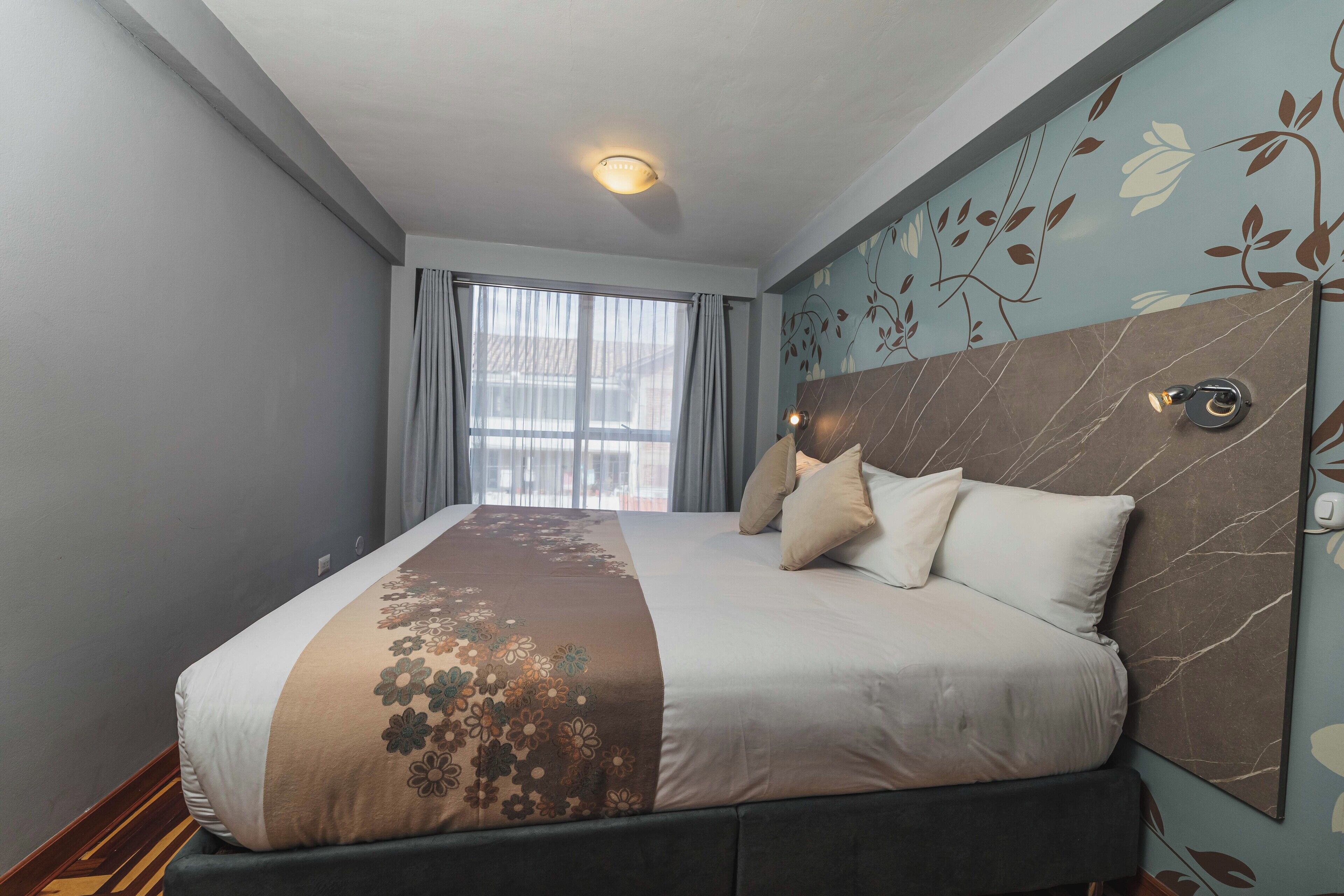 Habitación de hotel con una cama grande, cabecera con diseño floral, ventana con cortinas y una lámpara en el techo.