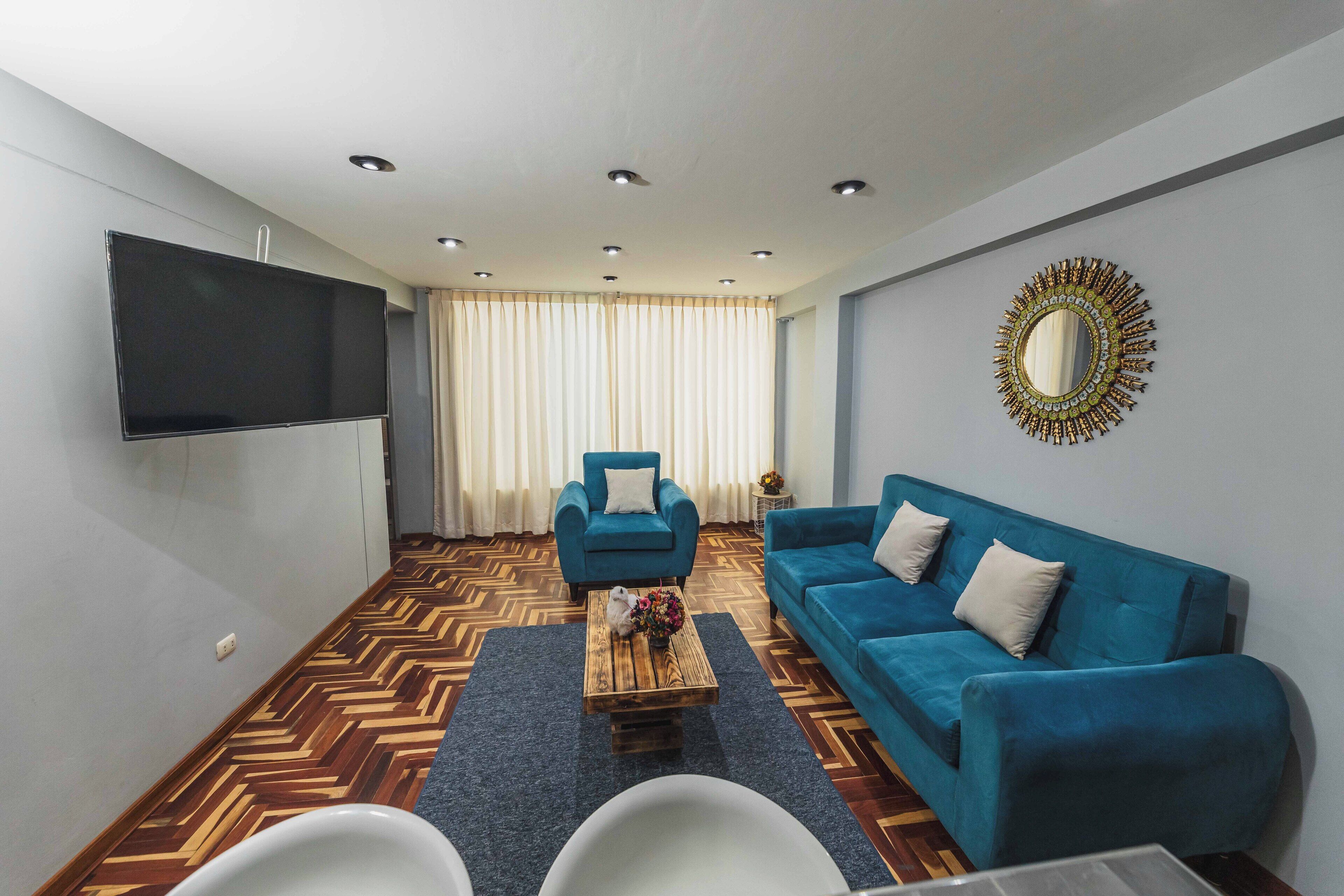 Un salon moderne avec un canapé bleu, un fauteuil assorti, une table basse en bois agrémentée de fleurs, un téléviseur à écran plat et un miroir décoratif accroché au mur.