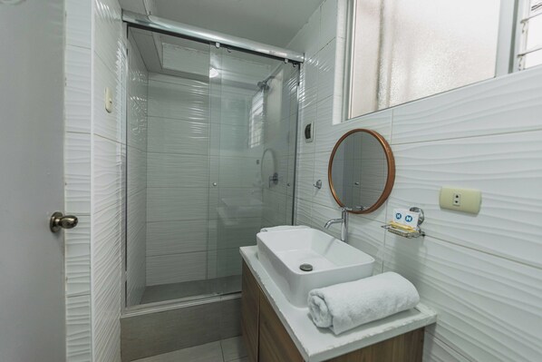 A bathroom with a glass-enclosed shower, a white sink with a wooden vanity, and a mirror with a wooden frame. - San Pedro Apartments (Cusco)