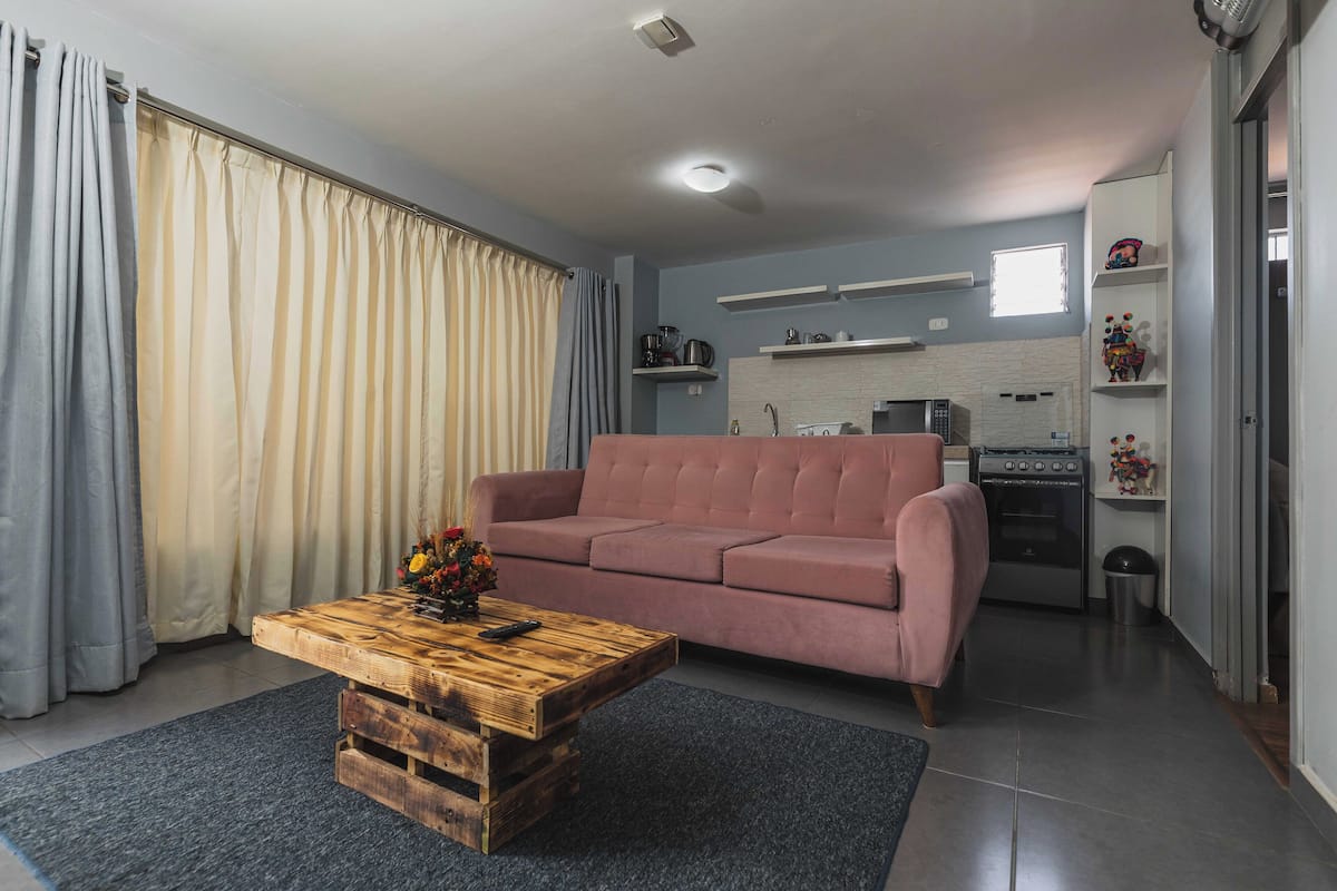 A living room with a pink sofa, a wooden coffee table, and a kitchen in the background.