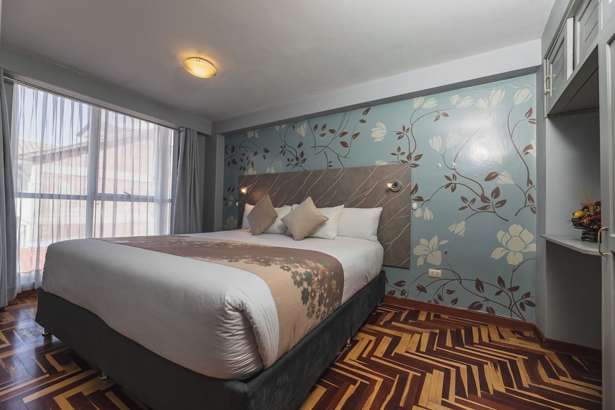 A hotel room with a large bed, a headboard with a floral design, a wooden floor, and a window with sheer curtains.