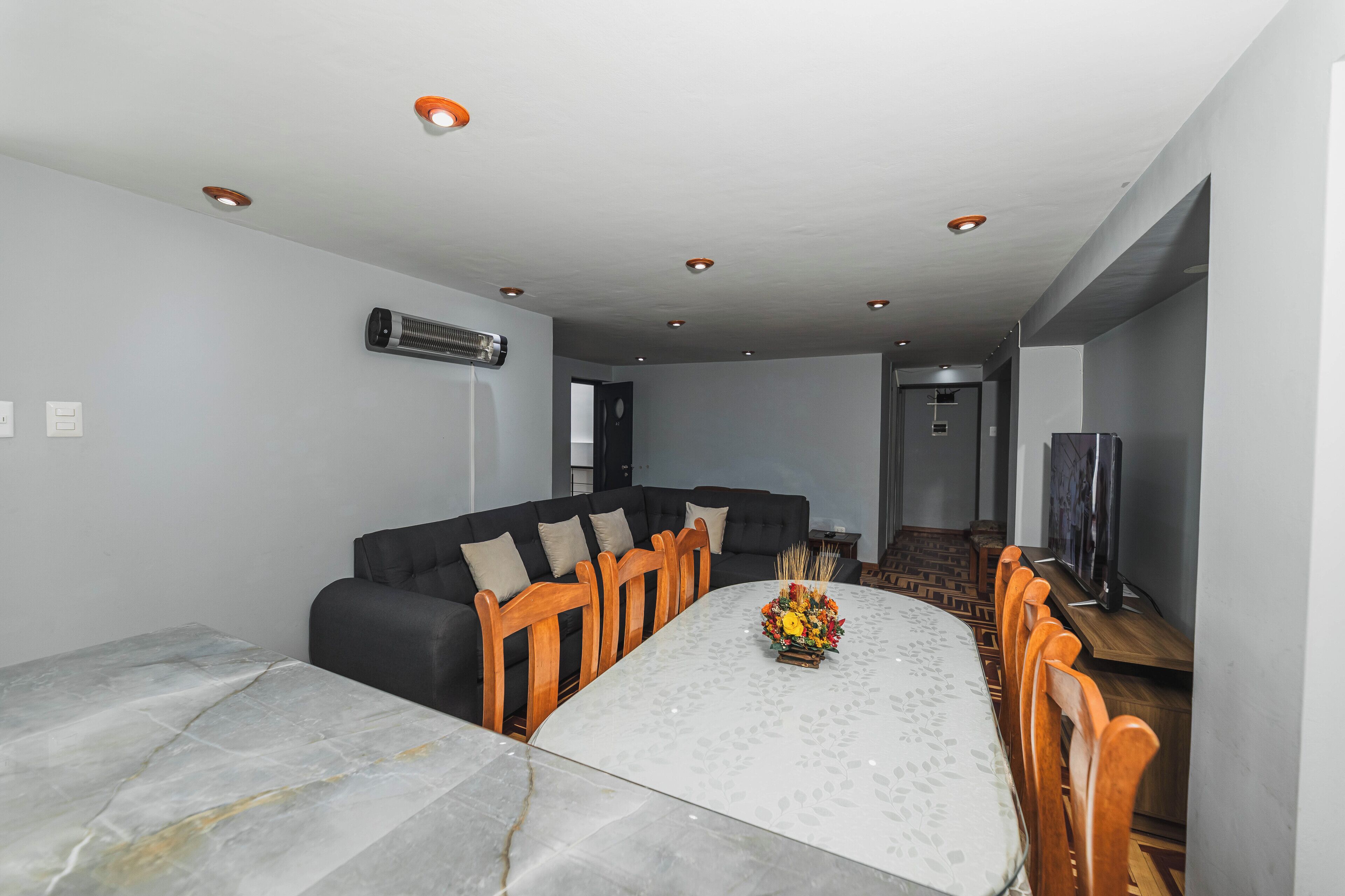Uma área de refeições moderna com mesa de mármore, cadeiras de madeira, sofá preto e iluminação embutida no teto.