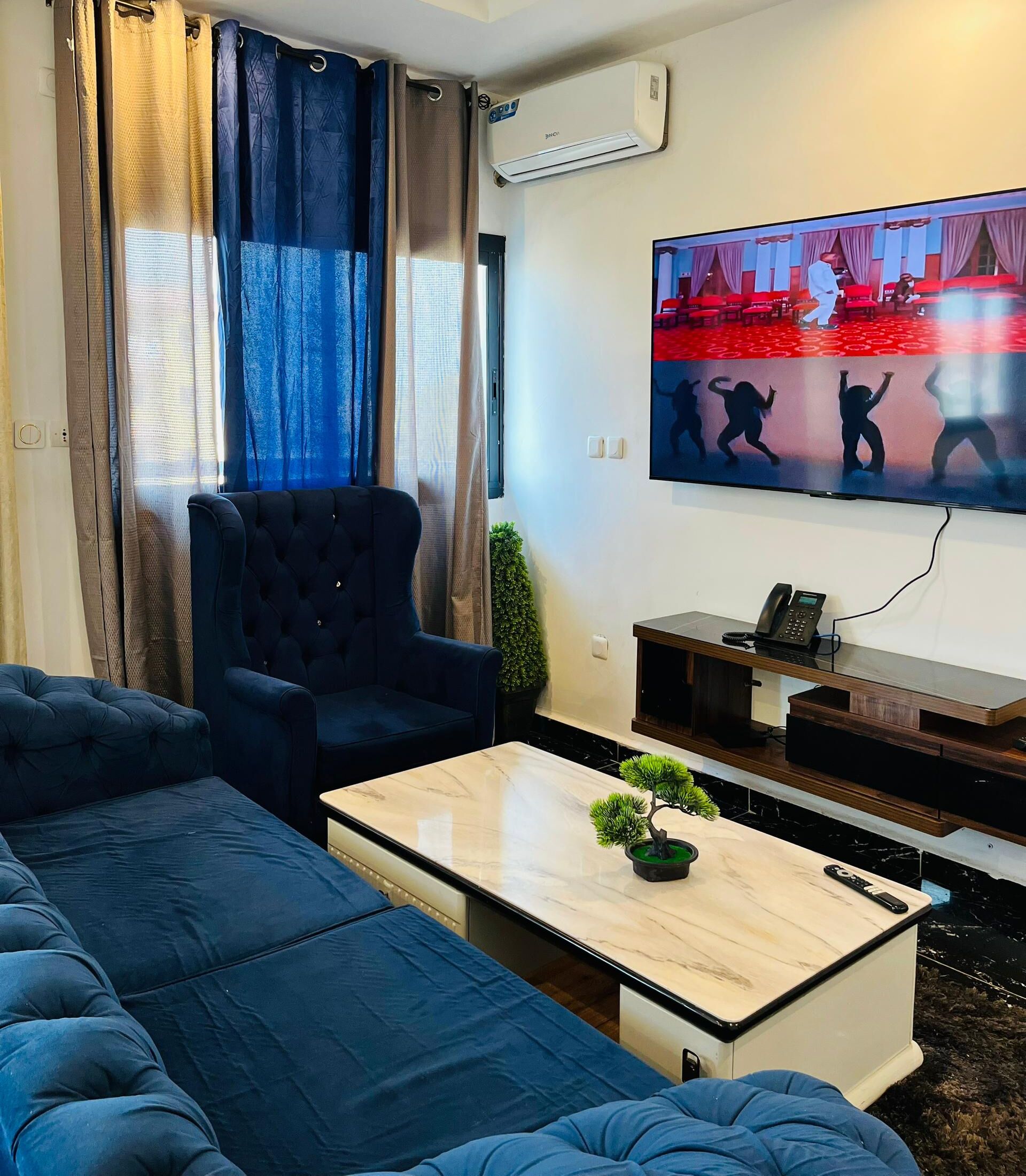 Een woonkamer met een grote bank, een donkere fauteuil, een salontafel met een plant, een aan de muur bevestigde televisie en een telefoon op een standaard.