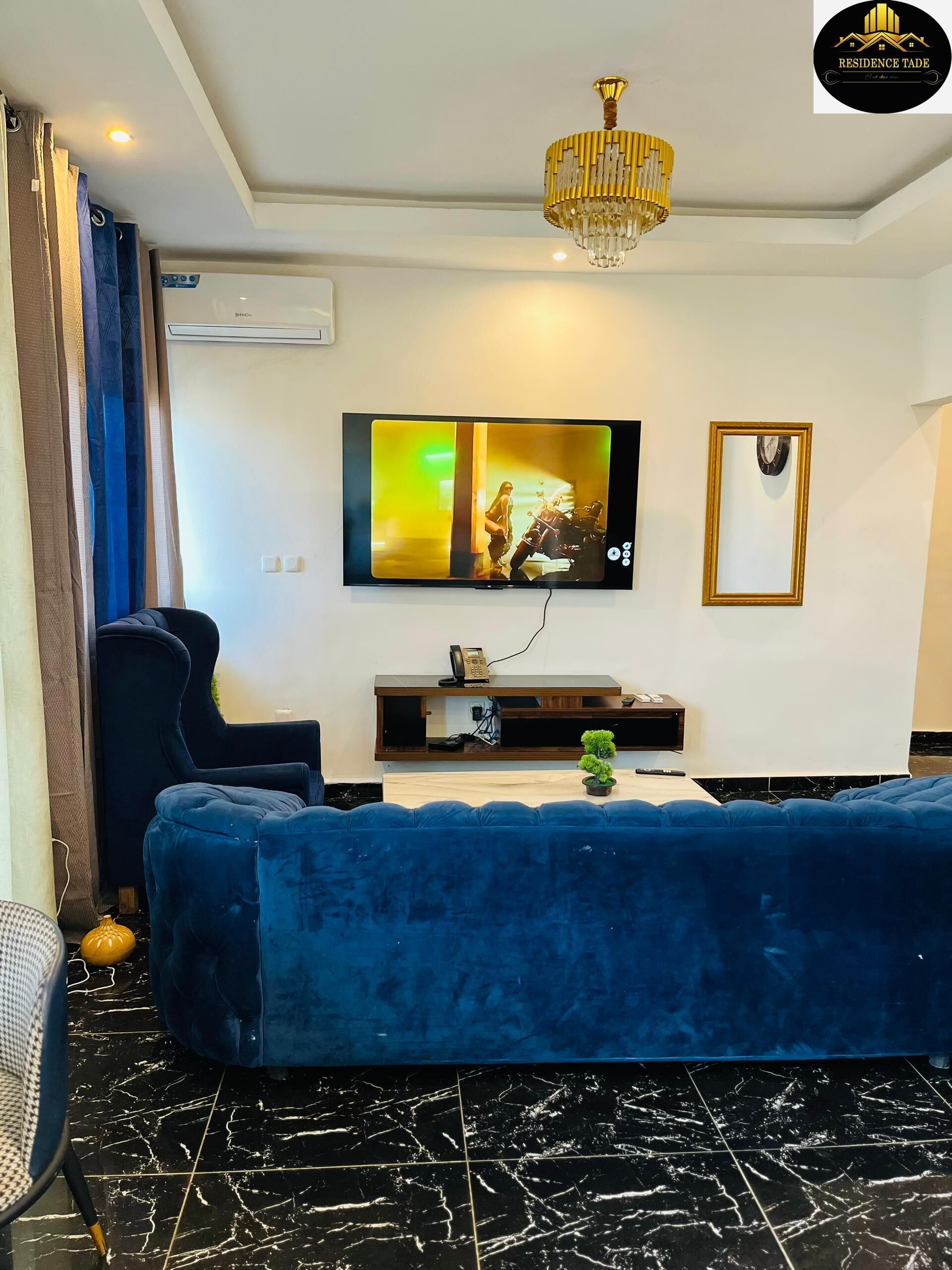Een moderne woonkamer met een blauwe bank, een wandgemonteerde televisie, een kroonluchter en een kleine plant op een houten console tafel.