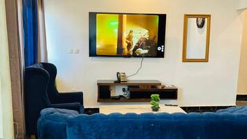 Een moderne woonkamer met een blauwe bank, een wandgemonteerde televisie, een kroonluchter en een kleine plant op een houten console tafel.