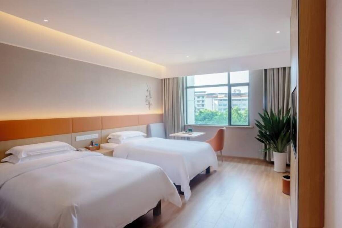 A hotel room with two beds, a small table, a chair, and a large window with a view of buildings.
