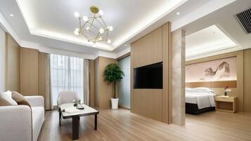 A modern hotel room with a wooden floor, a sofa, a table with a plant, a TV, and a bed with a nightstand.