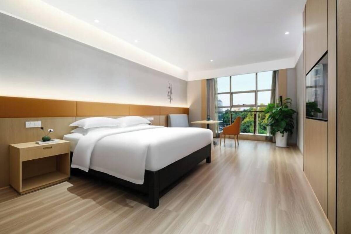 A modern hotel room with a large bed, bedside tables, a small table, a chair, and a large window with a view.