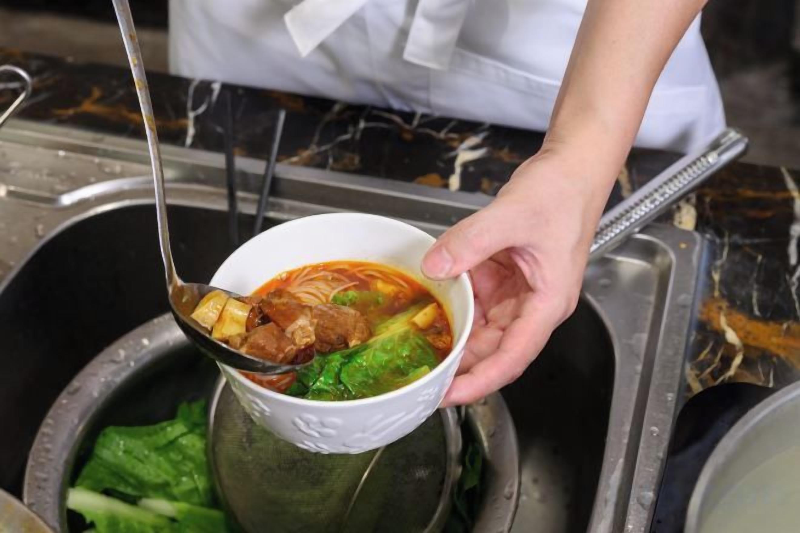 A person holding a bowl of soup with a spoon, near a sink.