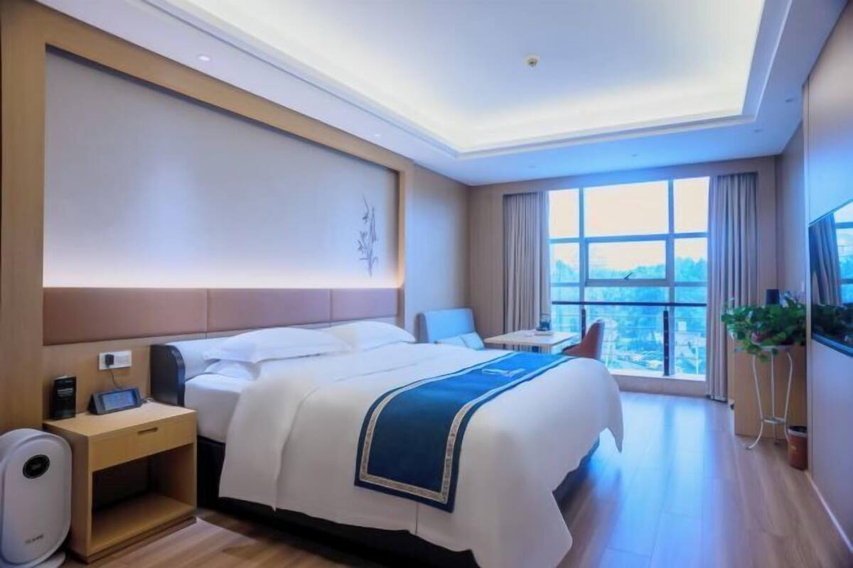 A hotel room with a large bed, bedside table, a small desk, a chair, a TV, and a large window with a city view.