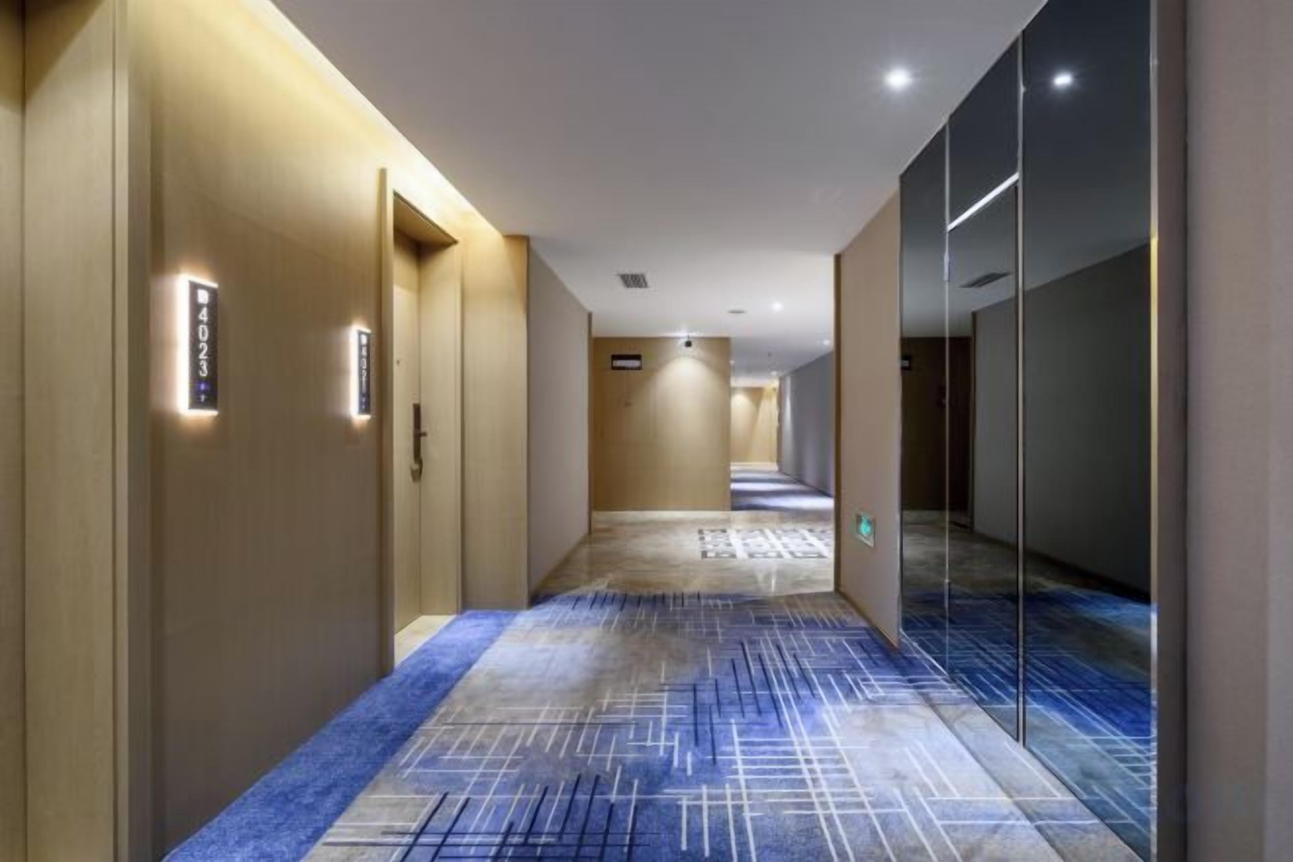 A hotel corridor with a carpeted floor, doors on the left, and a glass wall on the right.