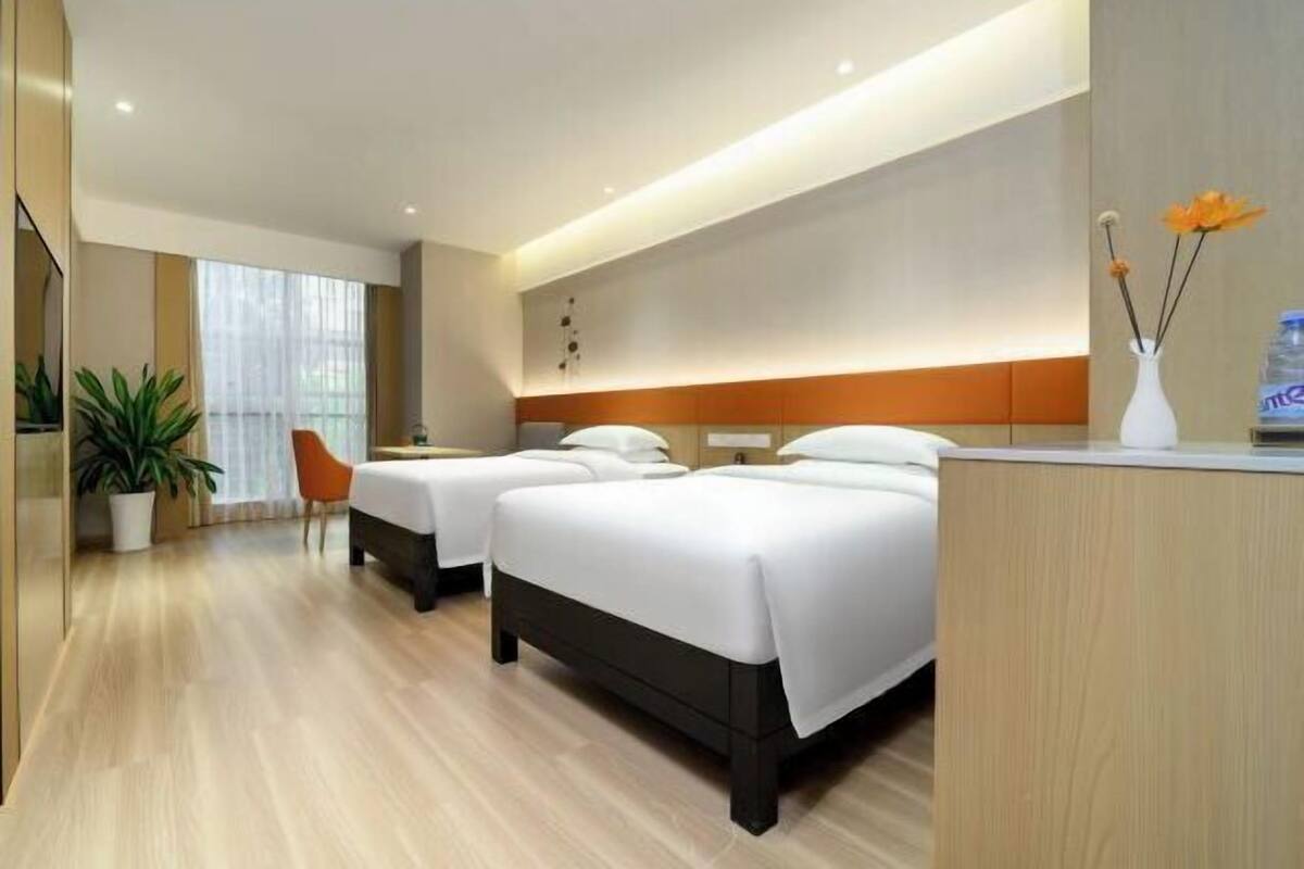 A hotel room with two beds, a desk, a potted plant, and a window with curtains.