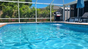 A clear blue pool with a tiled edge, surrounded by lush greenery and a covered patio area with outdoor furniture.