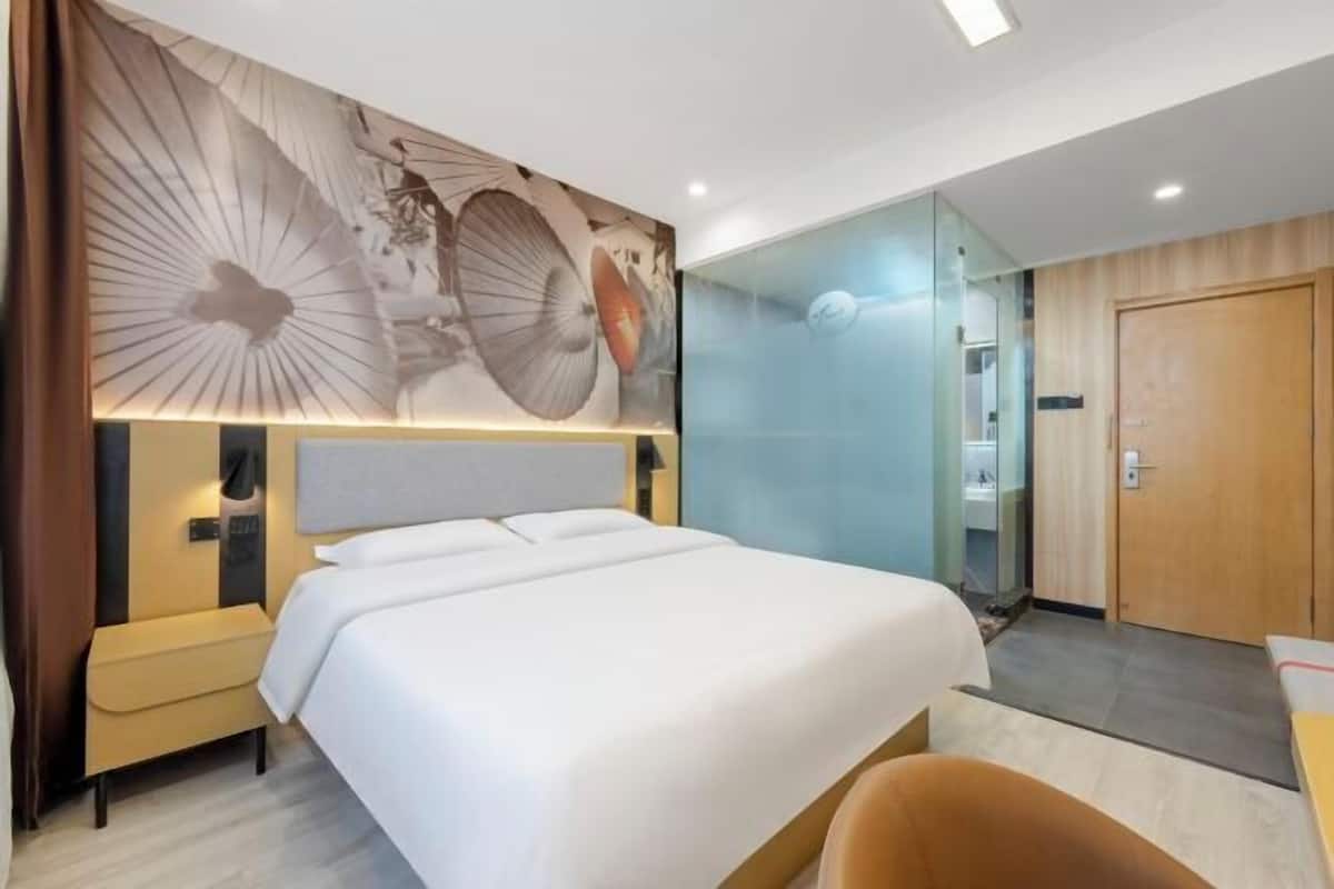 Una habitación de hotel con una cama grande, una mesita de noche, una silla, un separador de vidrio y una puerta de madera.