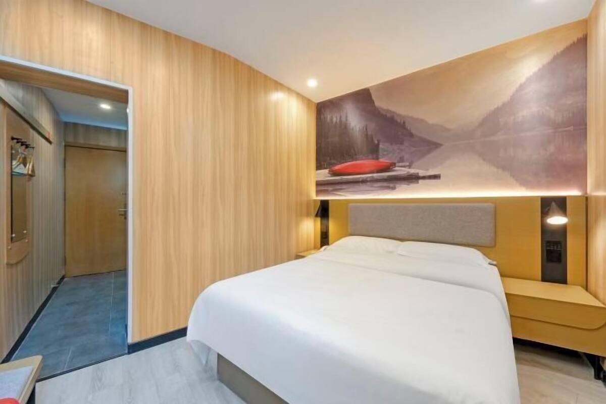 Una habitación de hotel con una cama, un armario de madera, un cuadro de un paisaje montañoso y una almohada roja.