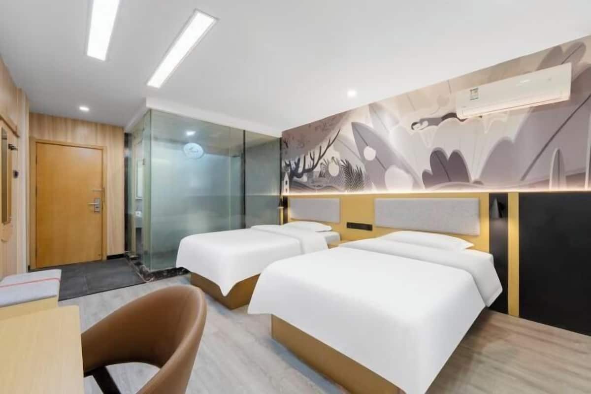 Habitación de hotel con dos camas, un escritorio, una silla, cabina de ducha de vidrio y una pared con arte abstracto.