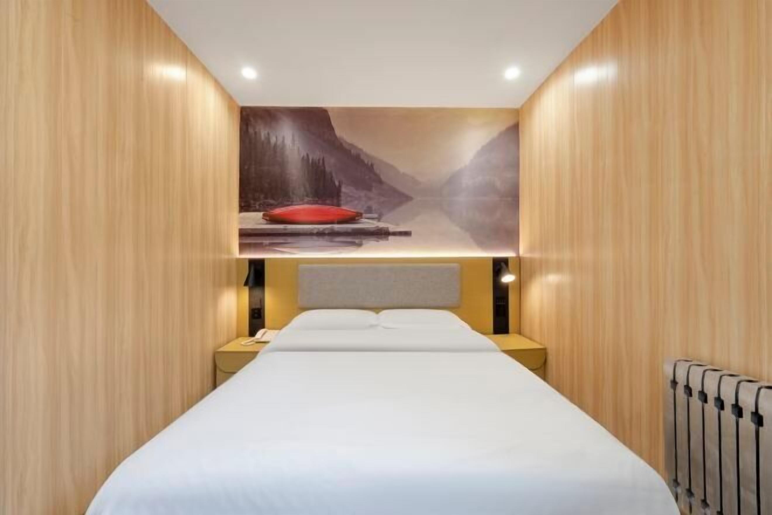 Ein Hotelzimmer mit Bett, Nachttisch, rotem Kopfkissen und Wandgemälde einer Berglandschaft.