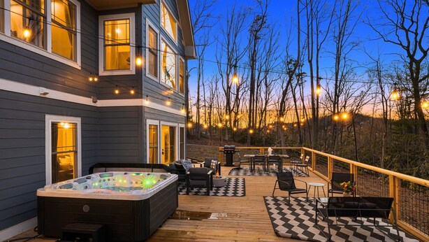 A deck with a hot tub, outdoor furniture, and a view of trees and a building. - 4k SqFt, Massive Deck , Games, Spa, Sauna, Firepit (Hendersonville)