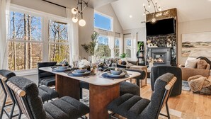 A spacious living area with a large dining table, comfortable seating, a fireplace, and large windows offering a view of trees. - 4k SqFt, Massive Deck , Games, Spa, Sauna, Firepit (Hendersonville)