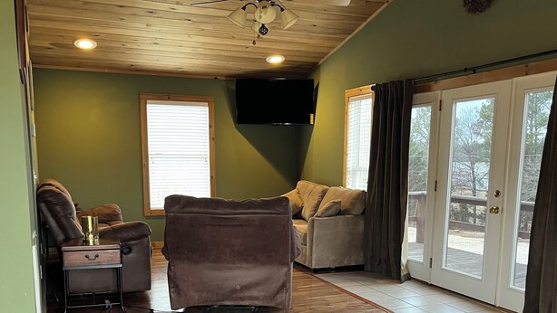 A spacious living room with a wooden ceiling, a ceiling fan, a flat-screen TV, and a large sliding glass door leading to an outdoor area. - Charming 1-bedroom house in Paris, TN. (Paris)