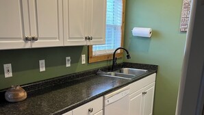 A kitchen with white cabinets, a black countertop, a sink, and a window with blinds. - Charming 1-bedroom house in Paris, TN. (Paris)