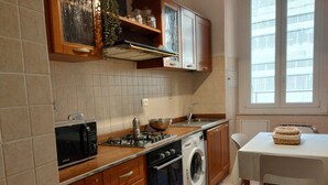 Cocina compacta con microondas, estufa, pileta y lavadora. Hay una mesa con sillas y una ventana con persianas.