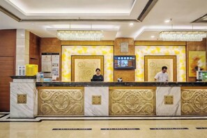 Ein Hotelrezeptionstresen mit Marmorplatte, zwei Rezeptionisten, einem Fernsehbildschirm und einer Chandelüster.