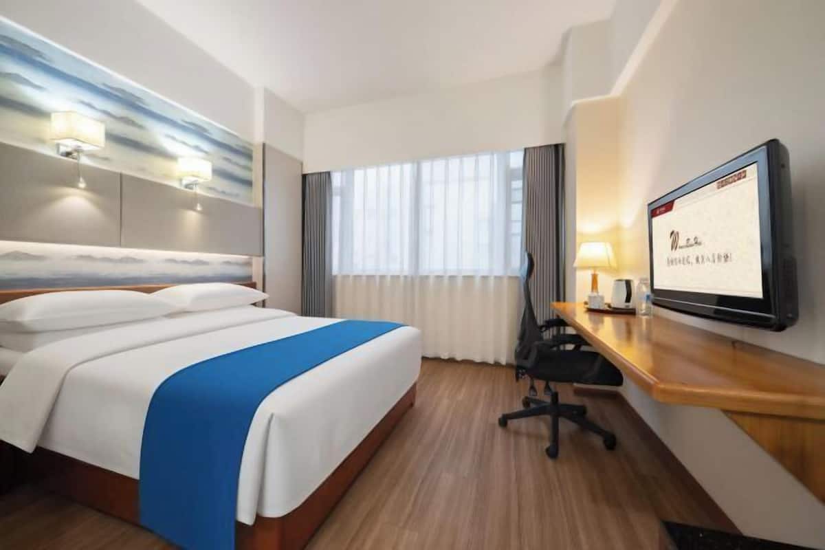 A hotel room with a bed, a desk, a chair, a TV, and a window with curtains.