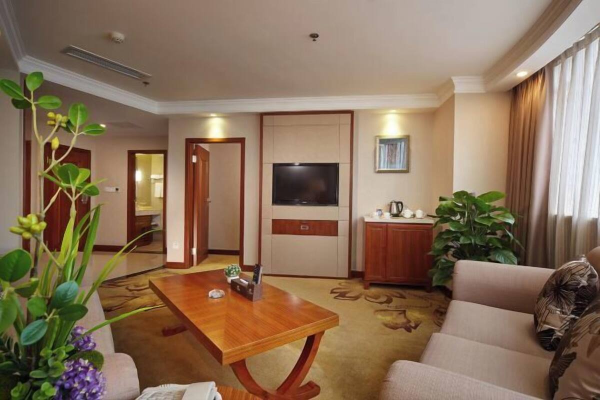 A hotel room with a wooden coffee table, a sofa, a TV, a cabinet, and a plant.