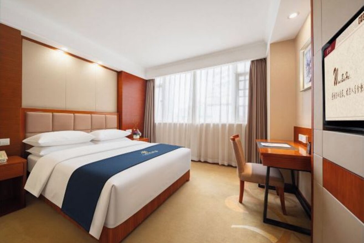 A hotel room with a bed, a desk, a chair, a nightstand, and a window with curtains.