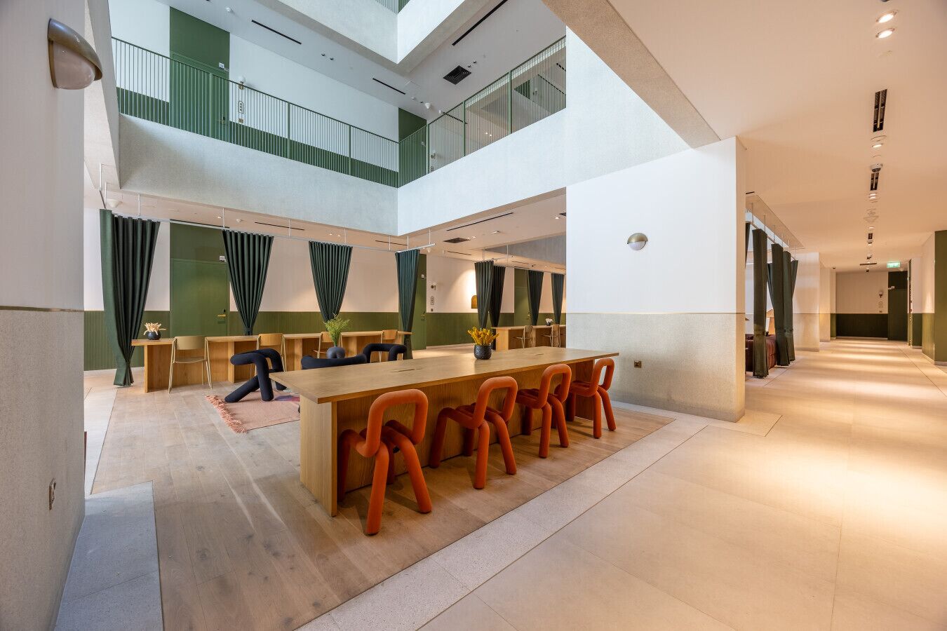 A spacious, modern interior with a central dining area featuring a long wooden table and orange chairs, surrounded by green curtains and wooden desks.