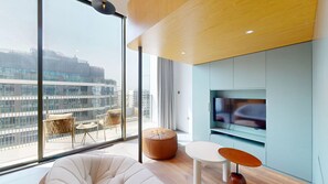 A modern living room with a large window, a flat-screen TV, and a seating area with a sofa and ottoman.
