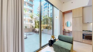 A modern apartment with a large sliding glass door leading to an outdoor patio, a green sofa, a kitchen with a built-in oven, and a view of high-rise buildings. - Mood 45 - Deluxe Studio: Chic with private Terrace (Jumeirah Garden City)