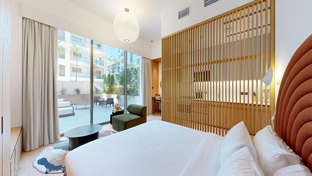 A modern hotel room with a large bed, a view of the outdoors, a wooden partition, and a dining area with a table and chairs. - Mood 45 - Deluxe Studio: Chic with private Terrace (Jumeirah Garden City)