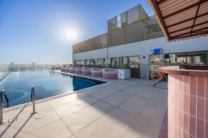A rooftop infinity pool with a clear view of the city skyline, a bar area with seating, and a sunshade structure.