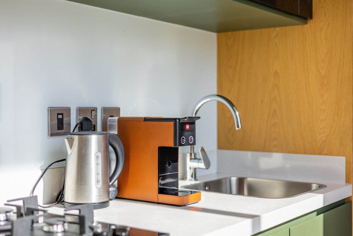 Eine moderne Küche mit einem Edelstahl-Wasserkocher, einer Kaffeemaschine und einer integrierten Espressomaschine auf einer weißen Arbeitsplatte.