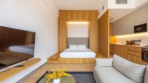 A modern hotel room with a bed, a TV, a sofa, and a kitchen area.