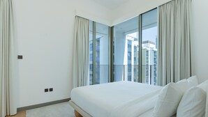 Une chambre d’hôtel moderne dotée d’un grand lit, offrant une vue sur la ville grâce à de larges fenêtres du sol au plafond, et disposant d’un balcon avec une balustrade.