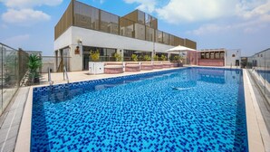 Una piscina con solárium empedrado, una zona de estar techada y un edificio moderno con amplios ventanales.