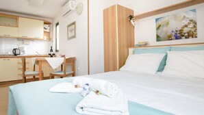 A hotel room with a bed, a desk with a chair, a kitchen area, and a painting on the wall. - Apartments Alba - Studio apartment (Rovinj)