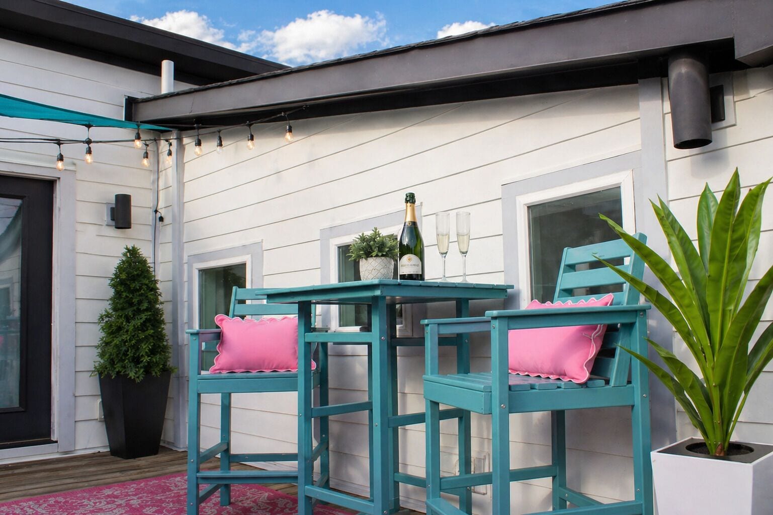Une terrasse avec deux chaises de couleur turquoise, une petite table, une bouteille de champagne et deux verres.