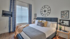 Une chambre à coucher avec un lit, des rideaux, une horloge murale, un téléviseur à écran plat, une table de chevet avec une lampe et une petite plante.