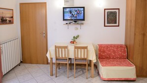 Een kleine woonkamer met een houten deur, een aan de muur bevestigde televisie, een tafel met twee stoelen, een rode bank en een lijst met een afbeelding aan de muur.