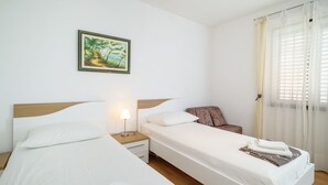 A hotel room with two beds, a nightstand with a lamp, a chair, a window with curtains, and a painting on the wall.