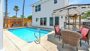 A swimming pool with a metal ladder, surrounded by a patio area with wicker chairs and a striped umbrella.
