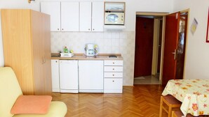 Eine kompakte Küche mit weißen Schränken, einer Mikrowelle und einer Kaffeemaschine. Ein Holztisch mit Blumenmuster bedecktem Tischläufer und zwei Stühle stehen daneben. Ein gelbes Sofa mit einem rosafarbenen Kissen ist im Vordergrund zu sehen.